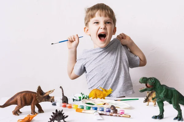 Hobby: o menino é um pré-escolar brincando e ri alegremente. Ele pinta figuras de dinossauros de brinquedo com tinta e um pincel . — Fotografia de Stock