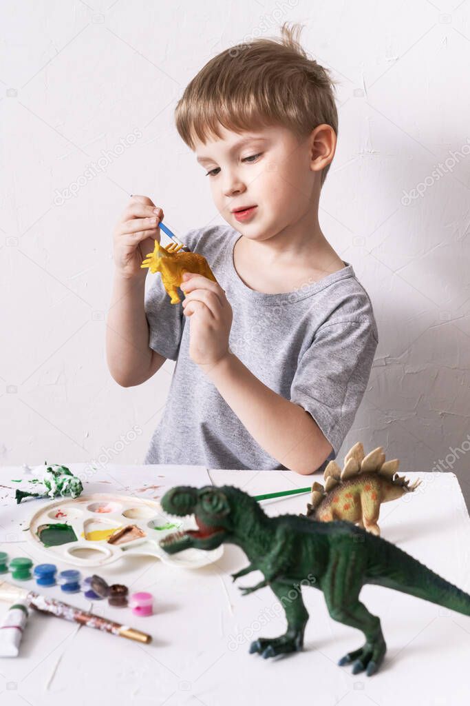 Hobby: a boy, a preschooler, paints with a brush and a tassel a small figure of a dinosaur toy.