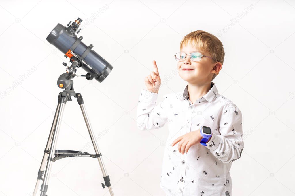 A little boy stands at a telescope and points his finger upwards.