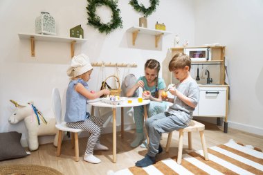 Easter holiday: children sit at the table and paint eggs with paints and brushes.
