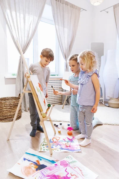 Talento: duas meninas em conjunto pintam em um cavalete com pinturas e uma escova em um grande quarto espaçoso . — Fotografia de Stock