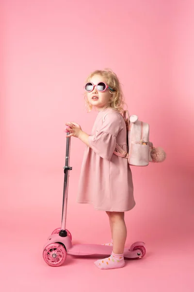 Una chica con gafas de sol y un vestido rosa con una mochila en la espalda rueda en una scooter rosa . — Foto de Stock