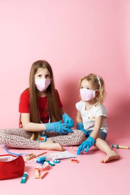 Kişisel koruyucu teçhizat: tıbbi maskeli ve eldivenli iki kız doktorculuk oynuyor ve oyuncaklardan tıbbi malzeme alıyor. Pembe arka planda bir portre.