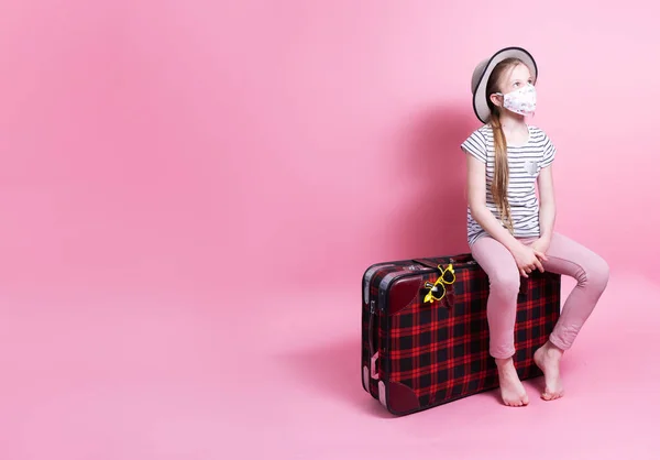 Podróż: Mała dziewczynka w słomkowym kapeluszu i masce medycznej siedzi na dużej czerwonej walizce i czeka na podróż. — Zdjęcie stockowe