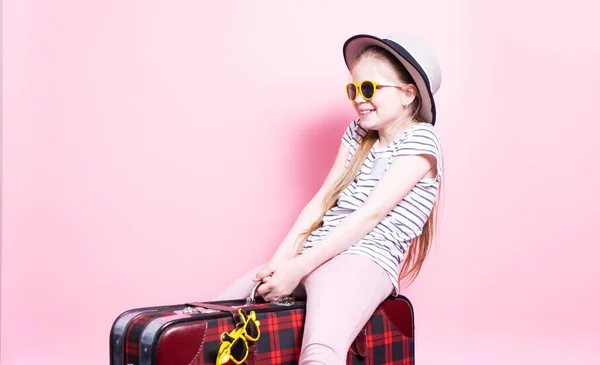 Podróże: Mała dziewczynka w słomkowym kapeluszu i okularach przeciwsłonecznych gra turystę i siedzi na dużej czerwonej walizce. — Zdjęcie stockowe