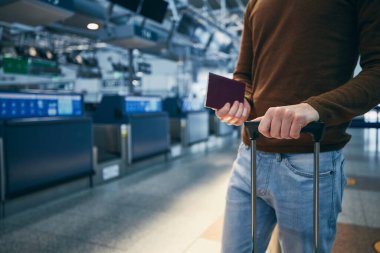 Passenger against check-in counter at airport. Hands of young man holding suitcase and passport.  clipart