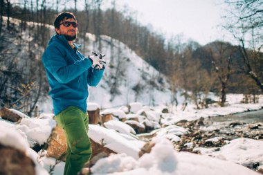 Genç erkek turist, dağlardaki bir nehrin kıyısında ısınıyor. Arka planda karla kaplı kıyılar ve ağaçlarla güzel bir kış manzarası. Tırmanma, yürüyüş, aktif yaşam, kamp konsepti.