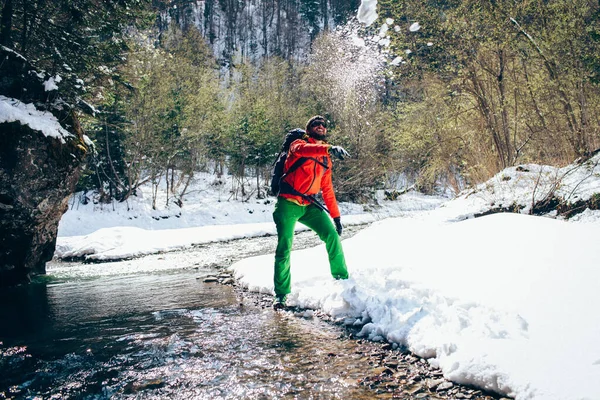 Young male tourist throws snow near a river in the mountains.Beautiful winter landscape with snow covered banks and trees  on background. Climbing trekking  and active life concept. Hiking equipment.