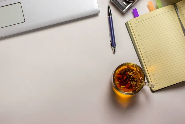Bir bardak bitkisel yatıştırıcı çay beyaz bir masanın üzerinde bir defter ve kalemin yanında duruyor. Evde çalışma kavramı..
