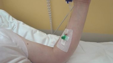 Hastanın koluna monte edilmiş venöz sonda görüntüsü. Hasta cihaza bakarak elini kaldırır, kamera el boyunca hareket eder ve ayrıntıları kapar..