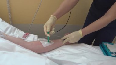 Hemşirenin elleri steril eldivenlerle venöz sondaya enjekte edilir. Hastanın elini arka plandaki usb-C konektöründe kateter, serum tüpü ve iki hemşire eli ile görebilirsiniz..