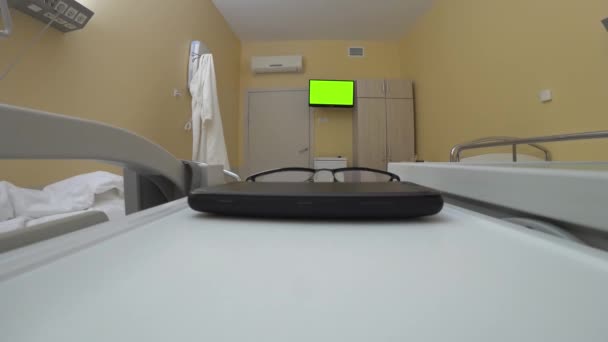 テーブルの上の空の医療病棟では スマートフォンや眼鏡 目に見えない患者の手は 金属のバーにスマートフォンの直腸キャンドルを置きます クロマキーグリーンのテレビ画面の背景に非常に近いです — ストック動画