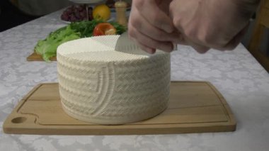 Taze ev yapımı keçi peynirinden büyük bir kafa kesmek. Beyaz peynirin desenli yüzeyi, arka planda taze sebzeler ve ahşap bir tahtanın sıcak yüzeyinde yatıyor..
