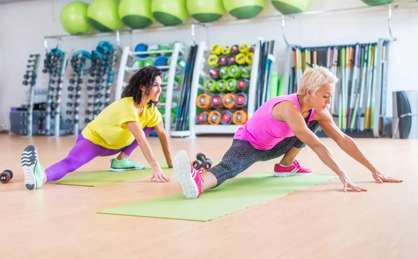 Seitenansicht von zwei Frauen beim Aufwärmen, die sich drinnen im Fitnesscenter auf Matten dehnen. Sportlerinnen beim Training in heller Sportbekleidung gegen bunte Geräte im Fitnessstudio — Stockfoto