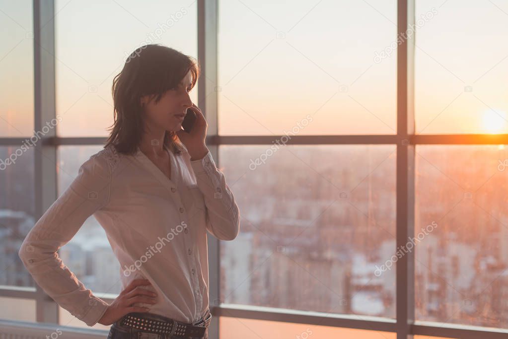 Young woman talking using cell phone at office in the evening. Female businesswoman concentrated, looking forward, calling by mobile device on her workplace.