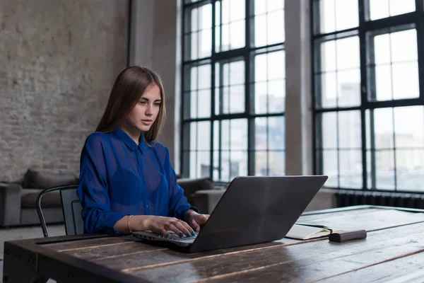 Muito jovem morena trabalhando no laptop na mesa de madeira no espaçoso estúdio escuro loft — Fotografia de Stock