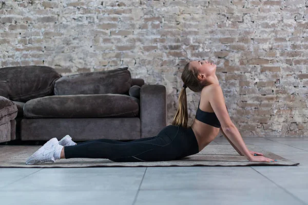 Jolie jeune femme en tenue de sport noire échauffant les muscles avant l'entraînement, étirant sa colonne vertébrale, faisant de l'exercice d'arc arrière dans un studio élégant — Photo