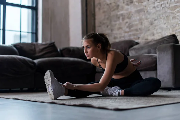 Mujer deportiva delgada en los músculos de calentamiento de ropa deportiva antes del entrenamiento haciendo ejercicio de estiramiento sentado en el suelo en casa —  Fotos de Stock