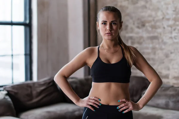 Porträt einer starken schlanken sportlichen kaukasischen jungen Sportlerin mit schwarzem Sport-BH, die mit den Händen auf den Hüften in die Kamera im Loft-Studio blickt — Stockfoto