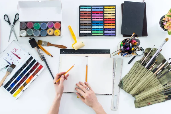 Artist drawing graphic sketch at sketchbook. Workplace, workspace. Top view photo of artistic tools lying on work-table: gouache, crayons palette and paintbrush collection. — Stock Photo, Image