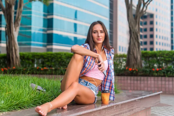 Gelukkig mooie jonge vrouw met blote benen zit op stenen beteugelen in het park en luisteren naar muziek in hoofdtelefoons. — Stockfoto