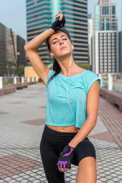 Retrato de jovem e cansada mulher fitness fazendo uma pausa do treino, limpando o suor da testa, ouvindo música. Atleta feminina descansando depois de se exercitar ao ar livre na rua da cidade — Fotografia de Stock