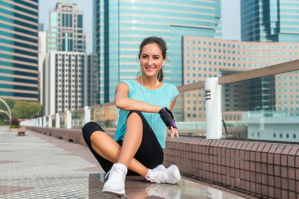 Chica fitness relajarse después de la sesión de entrenamiento sentado en el banco en el callejón de la ciudad. Mujer atlética joven tomando un descanso de correr, escuchando música en los auriculares y sonriendo . — Foto de Stock