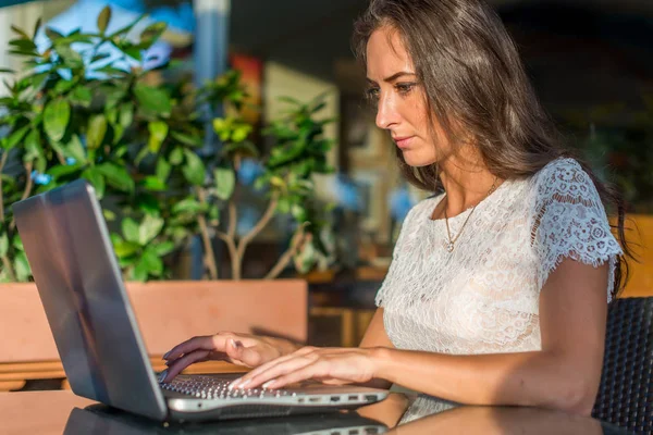 Sidovy av unga kvinnliga författare skriver på sin laptop när man sitter i park café. Flicka maskinskrivning netbook utomhus. — Stockfoto
