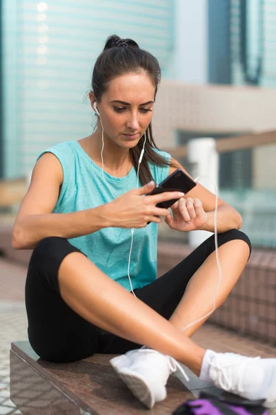 Ung sportig kvinna vila efter träningen med hjälp av sin smartphone och lyssna på musik i hörlurar. Idrottsman löpare i sportswear tar en paus — Stockfoto