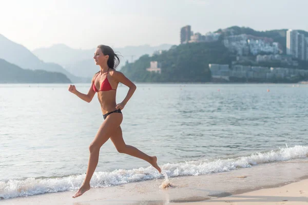 Passar kvinnliga idrottare bär bikini körs på stranden med solen skiner i kameran och hotel resort hills i bakgrunden — Stockfoto