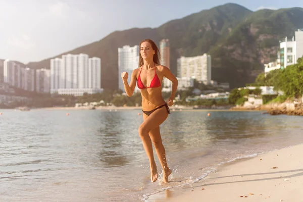 Fit athlète féminine portant un bikini courir sur la plage avec le soleil brille à la caméra et hôtel station collines en arrière-plan — Photo