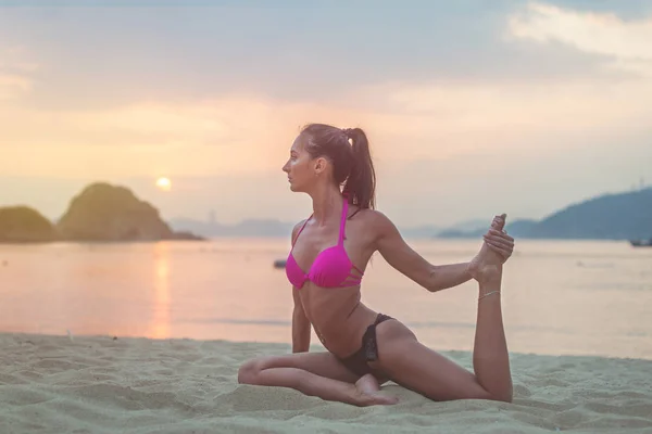 Mujer joven con sostén rosa y bragas negras sentadas en la playa estirando las piernas durante la puesta de sol en el mar. Chica de fitness haciendo ejercicios a la orilla del mar . —  Fotos de Stock