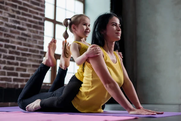 Mujer caucásica joven haciendo ejercicio de estiramiento para la columna vertebral junto con un niño sentado en su espalda en el gimnasio —  Fotos de Stock