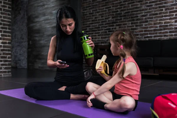Instructor de yoga para niños y una chica tomando un descanso después del entrenamiento sentada en la esterilla comiendo bocadillos, bebiendo agua, usando el teléfono en el gimnasio —  Fotos de Stock