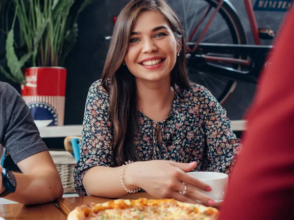 Retrato de una joven mujer bastante sonriente mirando a la cámara sentada en el restaurante bebiendo café y comiendo pizza con amigos —  Fotos de Stock