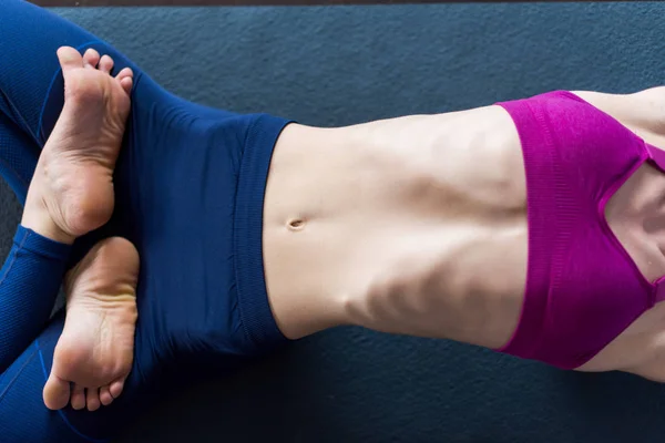 Close-up imagem do corpo feminino magro em sutiã esportivo e leggings deitado no tapete descalço fazendo backbend com pernas cruzadas na posição de lótus — Fotografia de Stock