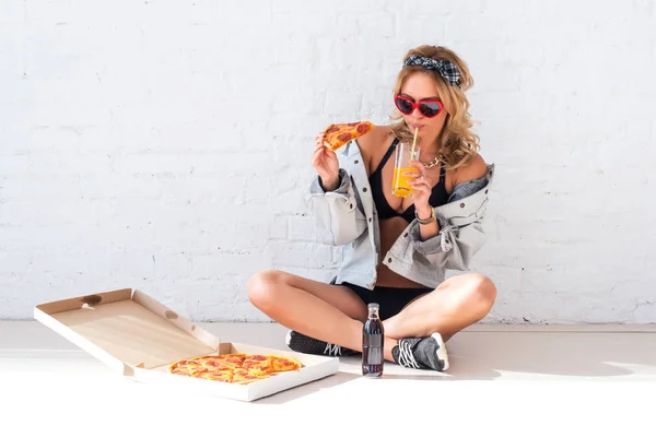 Ung kvinna äta en bit av pizza dricka saft som sitter på golvet bär solglasögon — Stockfoto