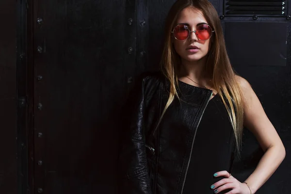 Foto de moda de modelo femenino atractivo en ropa de estilo rock agresivo y gafas de sol rosas posando en el interior oscuro — Foto de Stock