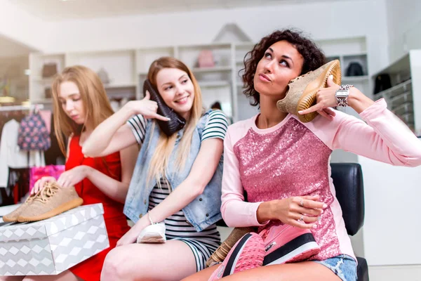 Tre giovani donne si divertono con le nuove calzature fingendo di fare una telefonata con le scarpe sedute in boutique — Foto Stock