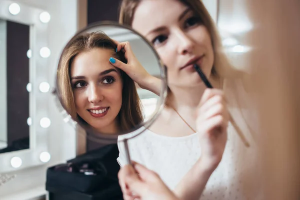 Joven cliente femenina mirando en el espejo mientras artista de maquillaje trabajando en sus cejas en el salón de belleza — Foto de Stock