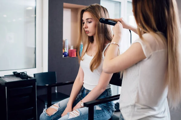 Hairdresser working on young girl s hairstyle curling her hair with straightening iron at hairdressing salon — Stock Photo, Image
