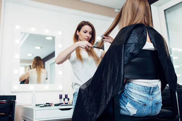 Young female hairdresser holding a strand combing girl s long fair hair in beauty shop — Stock Photo, Image