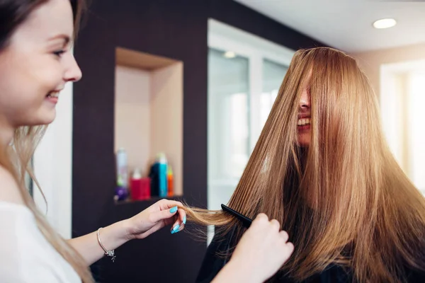 Kadın müşteri ile uzun düz sarı saçlar Kuaför Güzellik salonunda çalışan yüzünü önünde iplikçikleri dışarı penye süre gülüyor. — Stok fotoğraf