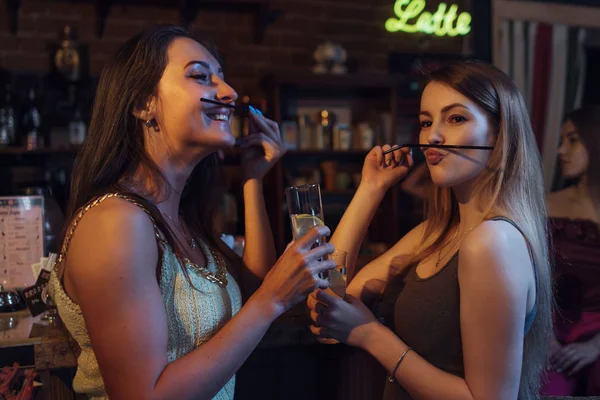 Girlfriends having a party holding glasses of cocktails, having fun, making moustache with straw in night club — Stock Photo, Image