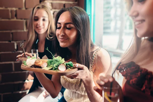 Gruppe von Mädchen beim Mittagessen in einem schicken Restaurant. Lächelnde junge Frau genießt den Duft von köstlichem Salat, der auf einem Holzteller serviert wird — Stockfoto