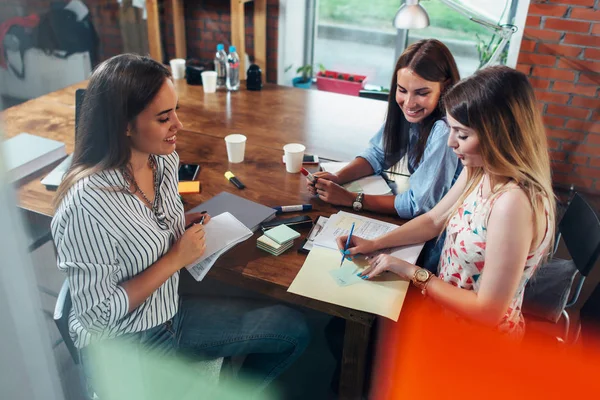 Grupp av leende kreativa kvinnor diskuterar ett projekt som sitter runt bordet att göra anteckningar i office — Stockfoto