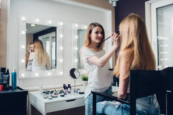 Kaukasische schoonheidsspecialiste dragen casual kleding die gebruikt worden voor de toepassing van make-up aan jonge vrouwelijke client in de schoonheidssalon — Stockfoto