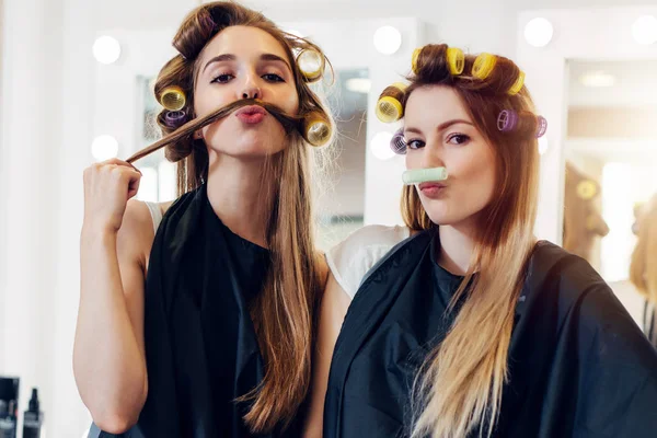 Två söta flickvänner i svart cape bär papiljotter gör moustache ur deras hår och roller i frisörsalong — Stockfoto