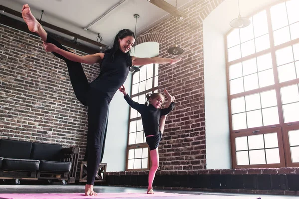 Sonriente madre y linda hijita en ropa deportiva negra haciendo ejercicio de estiramiento de pierna partida de pie manteniendo el equilibrio — Foto de Stock