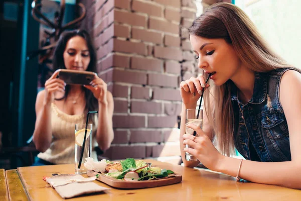 Two girlfriends having healthy lunch in cafe. Young woman taking picture of food with smartphone posting on social media — Stock Photo, Image